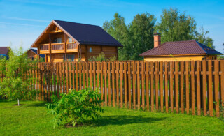 wooden fence on green grass with a bush and two houses in background